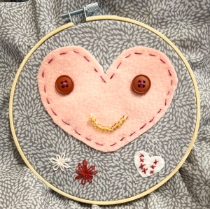 Pink embroidered heart with red button eyes and a gold embroidered smile.
